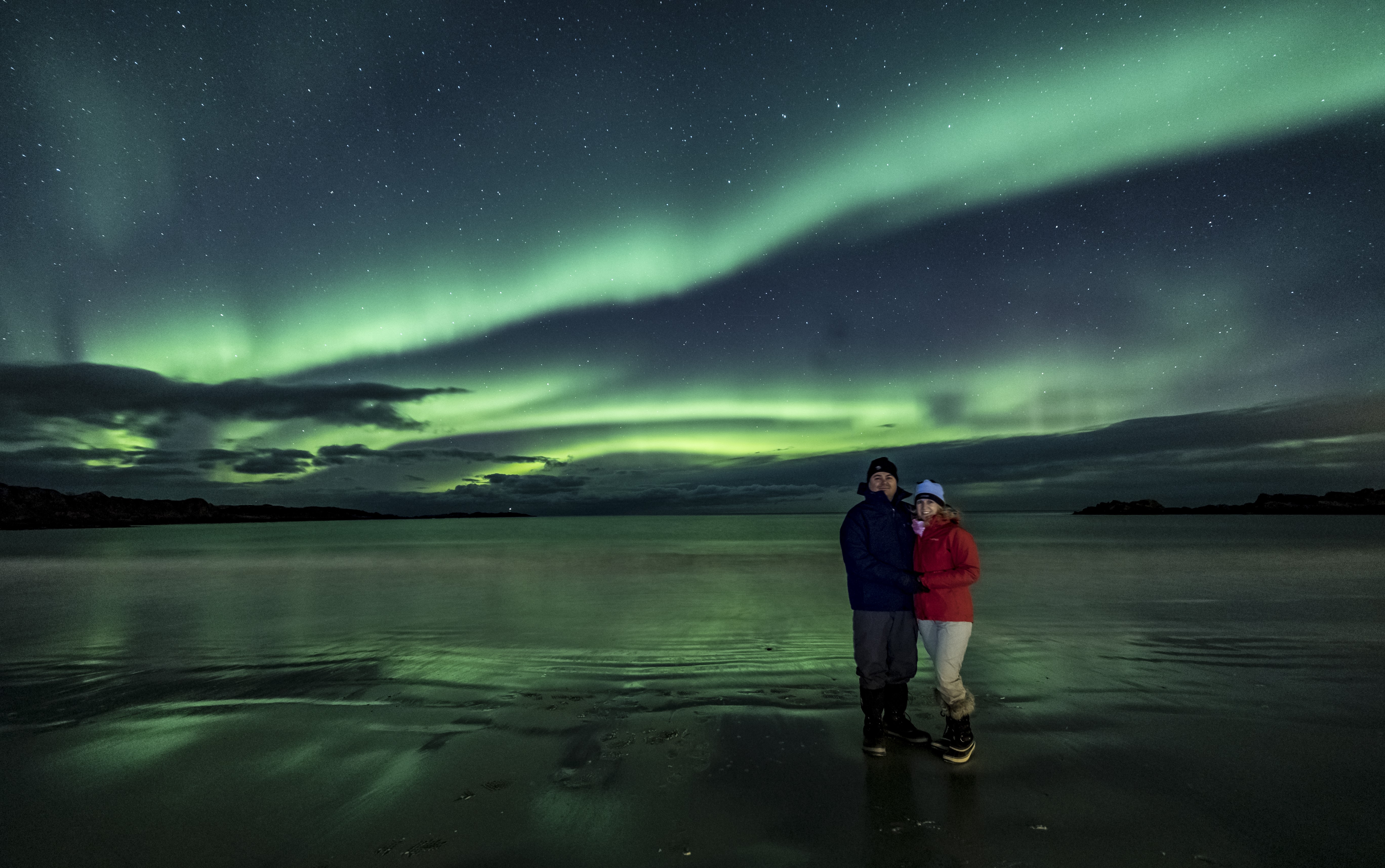 Two people standings on a beach with Aurora Borealis above reflecting into the lake.